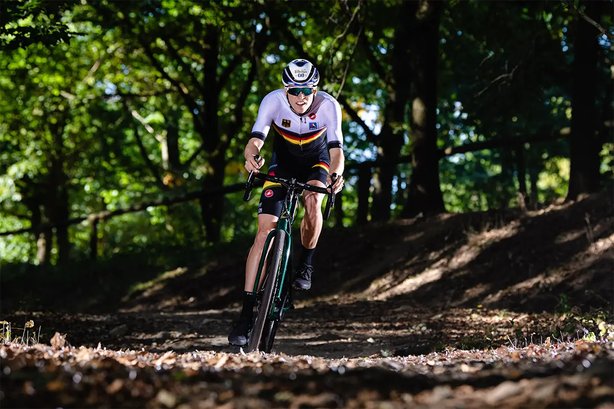 Gravelbike-Fahrer im Deutschlandtrikot fährt auf einem grünen Gravelbike durch einen Waldweg voller Laub.