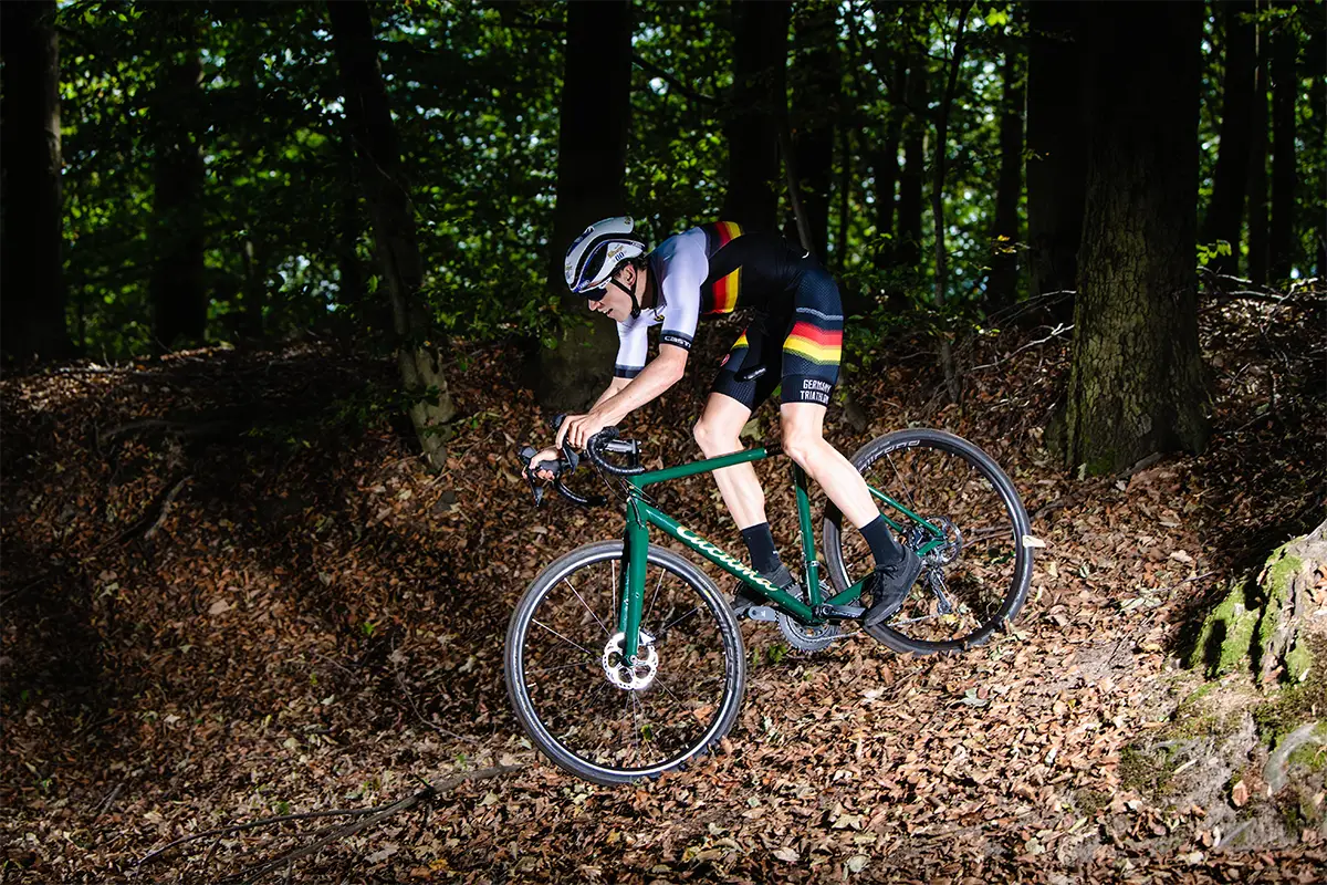 Gravelbike-Fahrer im Deutschlandtrikot fährt auf einem grünen Gravelbike eine Laubstrecke bergab.