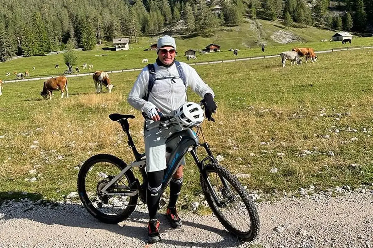 Radfahrer mit grauem E-Fully steht auf einer Wiese mit grasenden Kühen im Hintergrund.