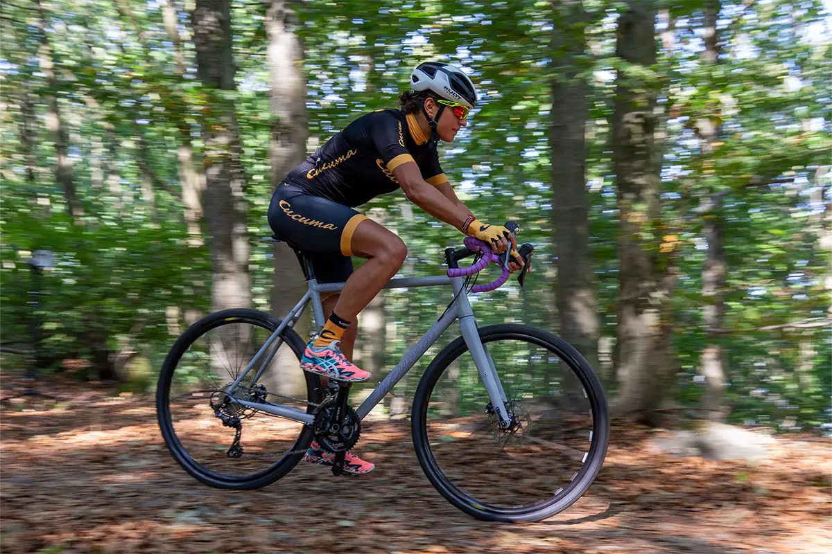 Gravelbike-Fahrerin auf grauem Gravelbike mit lila Lenkerband fährt durch einen Waldweg.